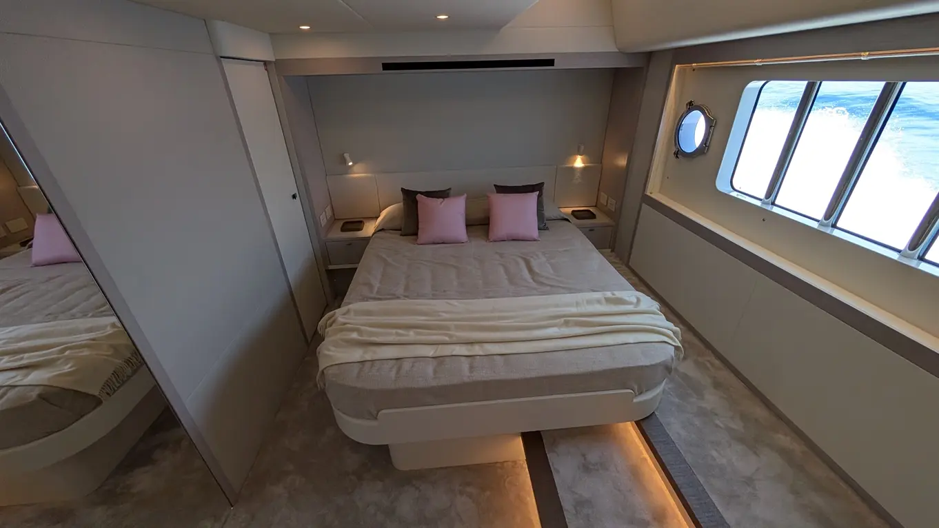 A modern yacht bedroom featuring a large bed with pink cushions, soft bedding, and a view through a round window.